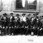 Image: Group of men and women sit and stand in two lines, most wearing dark clothing and hats