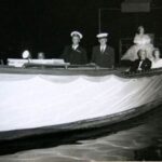 Image: a woman in an extravagant gown sits on a raised dias at the stern of a small boat. Other well dressed individuals sit along the sides of the boat.