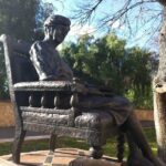 Image: A bronze statute of a middle-aged woman seated in a large chair. A stack of books is located next to the chair