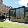 Image: The front of a modern, glass building with a multi-storey historic building in the background