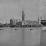 Image: A four-storey stone building is located adjacent to a waterfront wharf. Several ships are visible in the river in the foreground, and two large brick chimneys emerge from behind the stone building