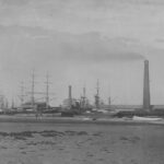 Image: A multi-storey stone building with large brick chimney sits near a river-front wharf. A larger chimney is located to the right of the building, and several ships are moored at the wharf