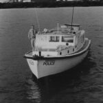 Image: A small motor boat with a lone man standing on its starboard side. A power station building is visible in the background