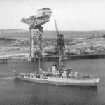 A warship gets underway in waters a short distance from a shore-based shipbuilding facility. The facility includes two large cranes and several buildings