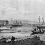 Image: A number of sailing ships are moored in a river. A cluster of buildings and wharves are visible in the background. A rowboat discharges people in the right foreground