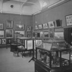 Image: A high-ceilinged room containing several pieces of artwork on its walls and a handful of display cases containing miscellaneous objects