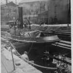 Image: A tugboat sits on stands in a dry-dock. A group of men sit facing the vessel in the foreground