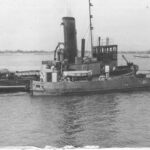 Image: A tugboat is moored against another a slightly smaller vessel in a river. A stretch of riverbank is visible in the background