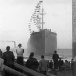 Image: A large iron ship a short distance from shore shortly after being launched. Several men on shore in the foreground look at the ship