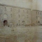 Image: A large, imposing three-storey nineteenth century warehouse stands near a port waterfront. Horse-drawn carts are positioned near large doors at the front of the building, and the masts of a sailing ship are visible in the distance