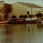 Image: A derelict tugboat is moored against a wharf next to a large complex of buildings. A number of other small boats are also moored at the same wharf