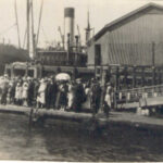 Image: A large group of people congregate on a floating dock next to a wharf. A steamship and warehouses are visible in the near background