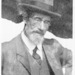 Image: A photographic portrait of an elderly Caucasian man wearing an Edwardian-era suit and hat. He has a white full beard and is wearing wire-rimmed spectacles