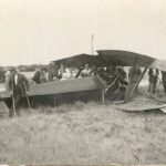 Image: A group of people examine the wreckage of a crashed biplane, including a man using crutches