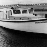 Image: A small motor boat idles along a waterfront. A warehouse is visible in the background