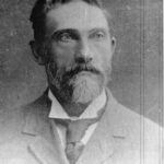 Image: Photographic head-and-shoulders portrait of a bearded, middle-aged Caucasian man. He has a full head of hair, light-coloured eyes and is wearing an Edwardian-era suit and tie