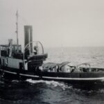 Image: A tugboat with the name ‘Yelta’ painted on its stern underway. The vague outline of a coast is visible in the distant background