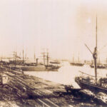 Image: A sailing ship is tied up alongside a long wharf. Horse-drawn carts full of cargo are positioned on the wharf next to the ship. Other sailing ships are visible in the background