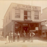 Image: A group of six men of various ages in Victorian attire stand in front of a shop. A painted banner on the top of the shop reads ‘Hy. Weman. Sail Maker & Ship Chandler’