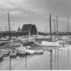 Image: A drawbridge within an active port is open to allow a vessel to pass beneath it. Several sail-boats and other small watercraft are moored in a boat basin in the foreground