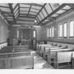 Image: The interior of a Christian chapel constructed from brick. It is an austere structure that features two lines of simple wooden pews and a small wooden altar