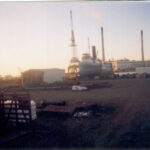 Image: A derelict tugboat in drydock at a boatyard. A large crane and a building with two tall chimneys is visible in the background