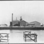 Image: A complex of multi-storey brick buildings and large, corrugated metal-clad warehouses fronted by a river. A large brick chimney is visible at one end of the complex