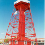 Image: An 1860s-era pre-fabricated lighthouse located along a modern city waterfront. The lighthouse tower is bright red and topped by a white lantern room. A chain-link fence surrounds the base of the lighthouse