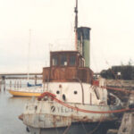 Image: A derelict tugboat is moored against a wharf. A handful of other small boats are also moored at the same wharf