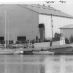 Image: A derelict tugboat is moored against a wharf next to a large complex of buildings. A number of other small boats are also moored at the same wharf