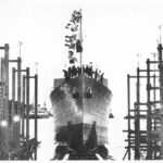 Image: A large iron warship is launched into the water. Several men stand on the bow of the ship, which is festooned in flags