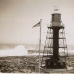 Image: A metal lighthouse stands on a desolate island next to a rough, wave-battered coastline. A roiling sea and high wind offer evidence of extremely poor weather conditions