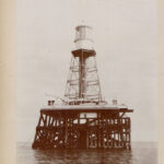 Image: A metal lighthouse with a large wooden support platform stands surrounded by water. A large buoy is visible in the distant background