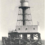 Image: A metal lighthouse with a large wooden support platform stands surrounded by water. The platform appears to be in a state of disrepair along one of its sides