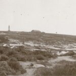 Image: A lighthouse stands at the top of a hill on a desolate island. A low stone building is present next to the lighthouse, and a wooden jetty along the shoreline protrudes into the ocean