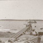 Image: A small wooden jetty projects into the ocean from a rocky coastline. A small steam vessel, four small sailboats and another island are visible in the background