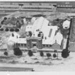 Image: An aerial photograph of a complex of buildings, including a large multi-storey brick structure built in mock-Tudor style. A railway station building is visible behind the complex