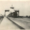 Image: A steam train passes beneath an observation tower on a bridge. A group of men in Edwardian attire perambulate along a walkway on the side of the bridge