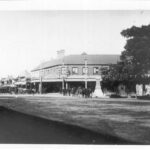 Image: The intersection of two dirt streets in a relatively large town. People dressed in early twentieth century attire walk along the street among several horse-drawn carts and carriages