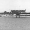 Image: The hull of an historic wooden sailing ship is towed across a modern bridge on a large flat-bed trailer. A number of modern boats and buildings are visible on the waterfront in the background