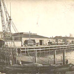 Image: A motor vessel gets underway from a wooden jetty. The waterfront of a port town is visible in the background