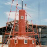 Image: The bottom section of a bright red, pre-fabricated 1860s-era lighthouse under construction. A large warehouse building is under construction in the immediate background