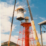 Image: A large crane hoists the lantern room of a pre-fabricated metal lighthouse to the top of the lighthouse tower. The lantern room is white and the lighthouse tower is red. A warehouse is visible in the background