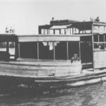 Image: A wooden-hulled motor boat transits between two points on a narrow river. The hull of the boat is in poor overall condition, and the vessel looks derelict