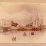 Image: Four rowboats sit unmoving in the middle of a river before a town waterfront. People within the boats are looking at the camera