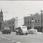 Image: Cars of 1950s vintage pass around a large plinth in the centre of an intersection in an urban area. The words ‘Keep Left’ are painted on the plinth, as are several large left-facing black arrows