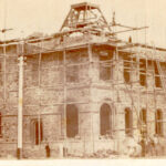 Image: An imposing, two-storey stone building under construction. The building is surrounded by scaffolding and a handful of men stand in front of its ground floor