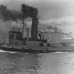 Image: A tugboat with ‘Yelta’ painted on its bow underway in an active port. A number of large ships and a wharf are visible in the background