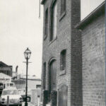 Image: A large, two-storey brick building with one large loading door in the centre of each floor. A hoist with block and tackle protrudes from the wall above the second storey loading door