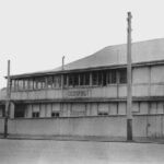 Image: Black and white photograph of a two storey building with a sign reading 'Cheer Up Hut' painted on the sign. A bag draped over the handlebars of a bicycle is just visible in the bottom left corner of the photograph.